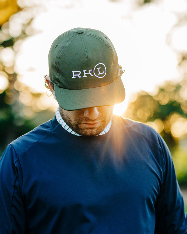 Man wearing a green cap with 'RK' logo, standing outdoors with sunlight filtering through.